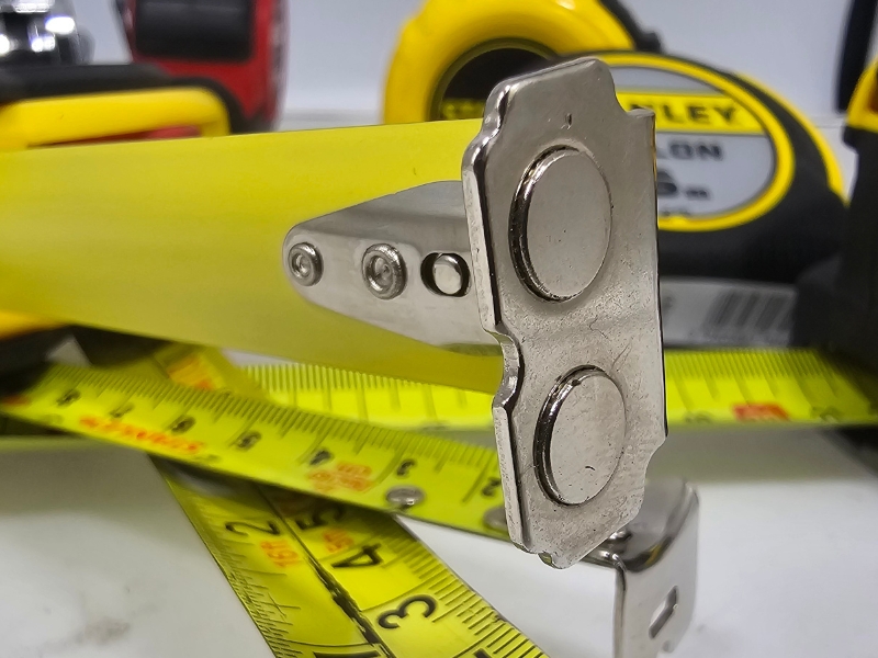 Duże zbliżenie stalowego zaczepu z wbudowanym magnesem na końcu taśmy miarki zwijanej, pokazujące szczegóły konstrukcji